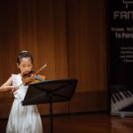 A young violinist performing from a music stand on the concert hall stage.