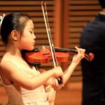 A young violinist mid-phrase, accompanied at the piano behind her.