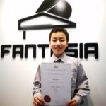 A student holding an AMEB Fifth Grade Piano certificate, awarded a Pass with High Distinction, in front of the Fantasia sign.