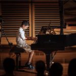 A boy playing a Steinway concert grand under stage lighting, watched by an audience.