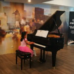 A young girl in a pink dress playing the grand piano at an in-house student concert.