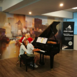 A teacher and a young student in Santa hats playing the grand piano together at a Christmas concert.
