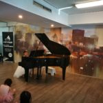 A small child performing at the studio grand piano while other children watch from the floor.